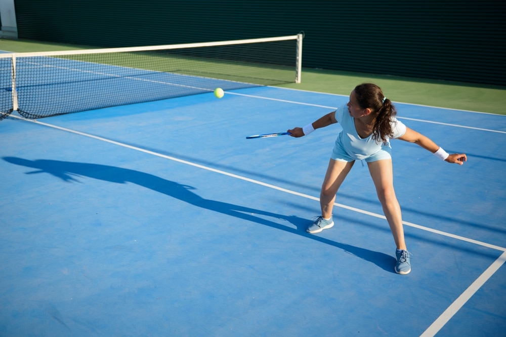 A young woman in motion hitting a tennis ball during a game on an outdoor blue tennis court, displaying agility and determination.